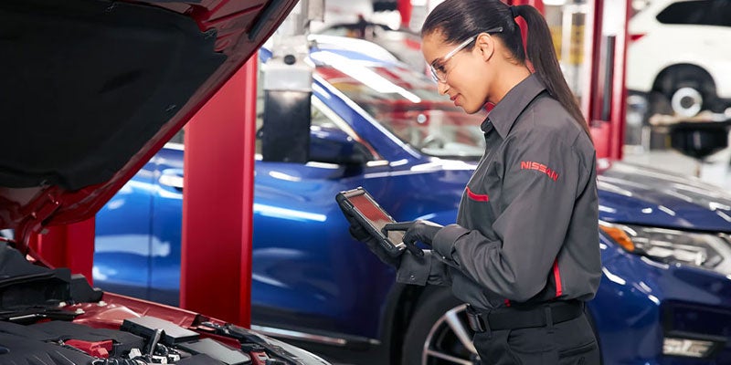 A woman at a car service center checks information on a tablet while standing next to a vehicle.