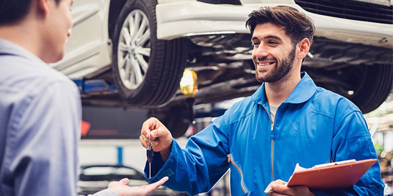 A man speaks with another man inside a busy car repair shop, surrounded by tools and vehicle parts.